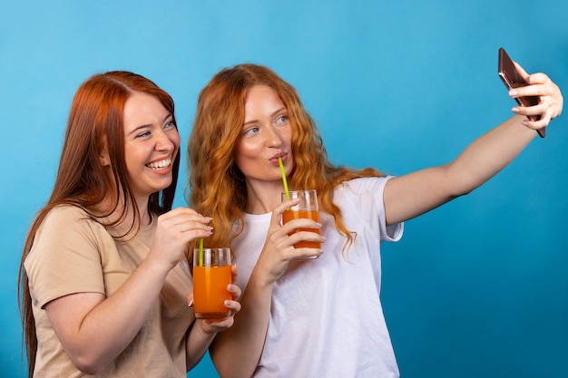 Freundinnen in Freizeitkleidung trinken Saft aus einem Strohhalm und machen Selfies. Auf blauer Wand isoliert. Menschen Lifestyle-Konzept.