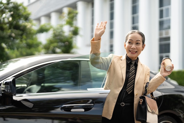 Freundliche asiatische Seniorin sagt hallo, winkt mit der Hand und begrüßt Sie mit einem glücklichen Lächeln, das ihr Luxusauto verlässt