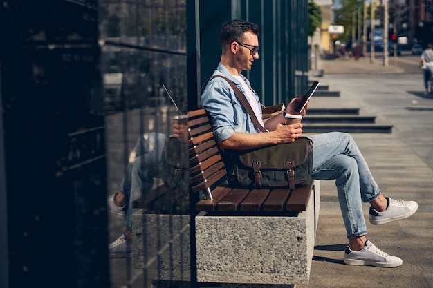 Freundliche brünette männliche person, die beim lesen der nachricht und beim kaffeetrinken ein lächeln auf dem gesicht behält
