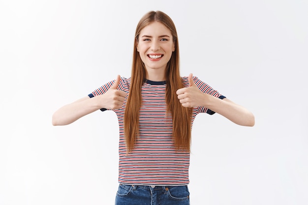 Freundliche, fröhliche, gut aussehende Kaukasierin in gestreiftem T-Shirt, die positive Antwort zeigt, Daumen hoch und zustimmend lächelt, akzeptierend nickt, wie eine tolle Idee, weißer Hintergrund stehend