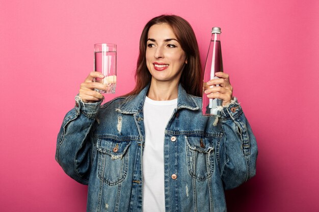 Freundliche junge Frau, die eine Flasche Wasser hält und ein Glas Wasser betrachtet