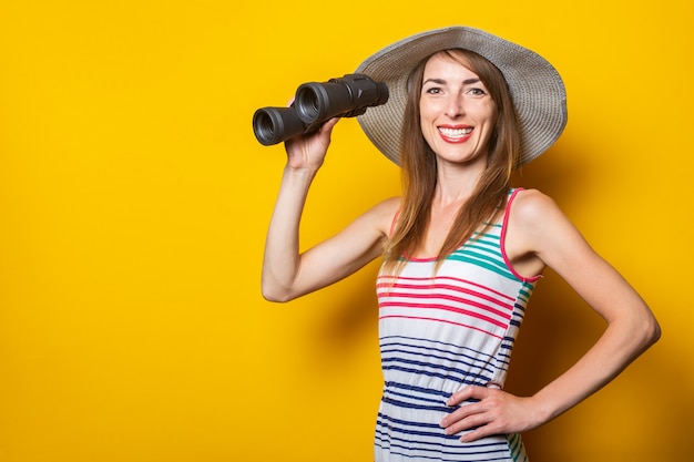 Freundliche junge lächelnde Frau in einem gestreiften Kleid hält Fernglas auf einem gelben Raum