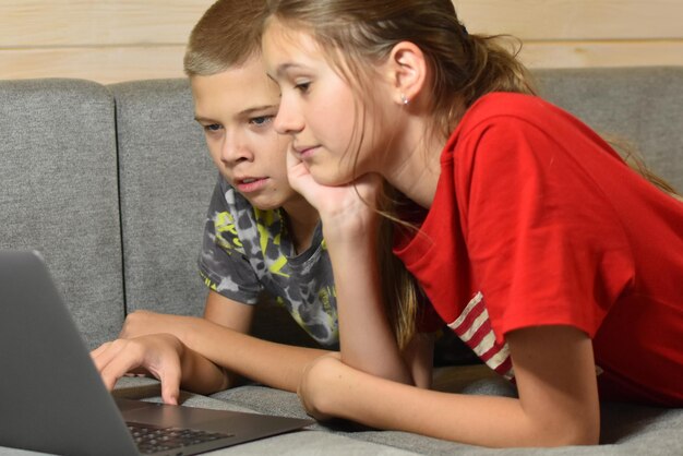 Freundliche Kinder liegen auf der Couch und schauen sich zu Hause Videos in einem Laptop an. Fernunterricht online von zu Hause aus