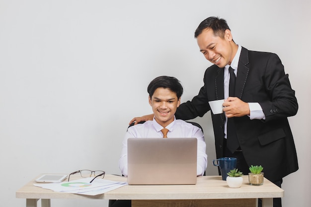 Freundlicher asiatischer Chef, der eine Tasse hält, und seine Mitarbeiter schultern, die am Schreibtisch arbeiten