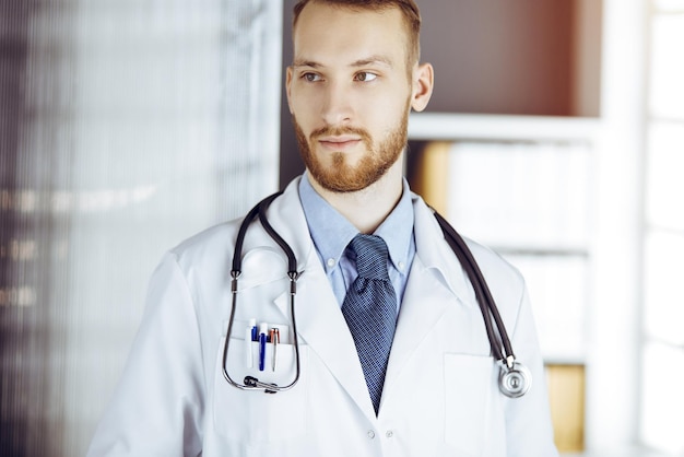 Foto freundlicher bärtiger arzt, der direkt in sonniger klinik steht. porträt des arztes. medizin-konzept.