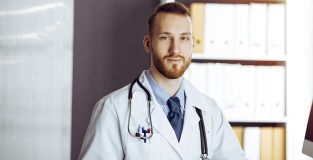 Freundlicher bärtiger Arzt, der in sonniger Klinik am Klemmbrett sitzt und schreibt. Medizin-Konzept.