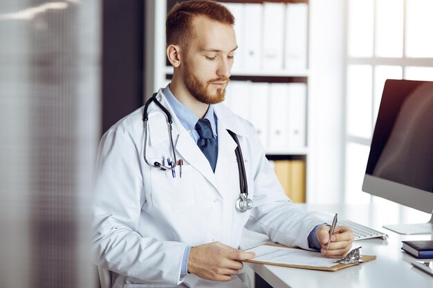 Freundlicher bärtiger Arzt und geduldige Frau diskutieren aktuelle Gesundheitsuntersuchung, während sie in sonniger Klinik sitzen. Medizin-Konzept.