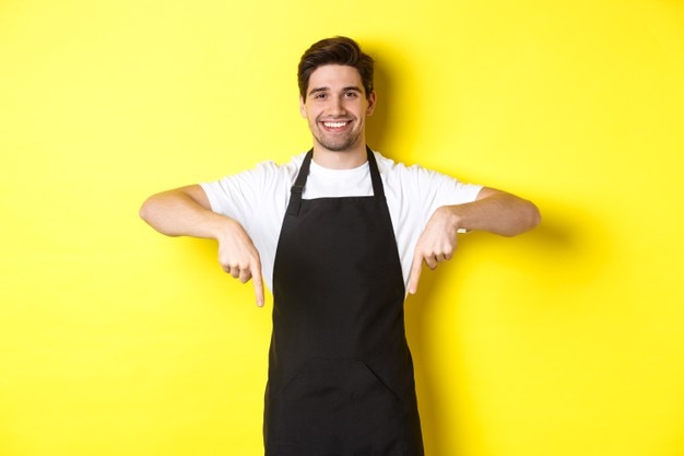 Freundlicher Barista in der schwarzen Schürze zeigt mit den Fingern nach unten und zeigt Ihr Logo-Banner, das über der gelben Wand steht