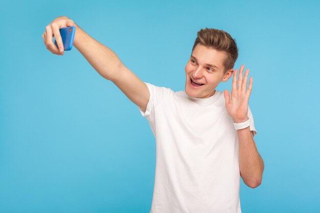 Freundlicher, glücklicher Mann in lässigem weißem T-Shirt, der Hallo sagt und Hallo sagt, während er über Videoanrufe spricht und ein Selfie mit dem Handy macht, das online chattet, Studioaufnahme isoliert auf blauem Hintergrund