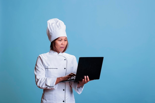 Freundlicher Koch, der kulinarische Rezepte auf dem Laptop durchsucht, die Zutatenliste überprüft und ein gesundes Abendessen kocht. Koch mit Restaurantuniform, der Gastronomiemahlzeiten im Studio mit blauem Hintergrund zubereitet