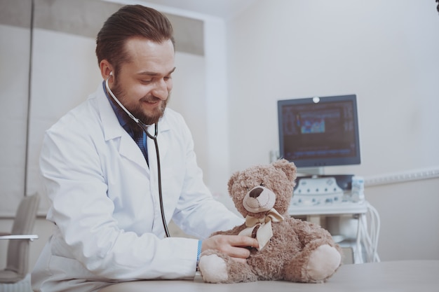 Freundlicher männlicher Kinderarzt, der in seiner Klinik arbeitet und vorgibt, Teddybärspielzeug zu untersuchen