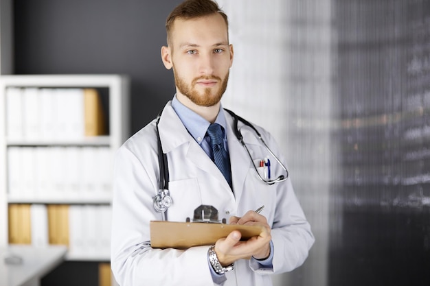 Freundlicher rotbärtiger Arzt steht und schreibt mit Klemmbrett in der Klinik an seinem Arbeitsplatz. Perfekte medizinische Versorgung im Krankenhaus. Medizin, Gesundheitswesen