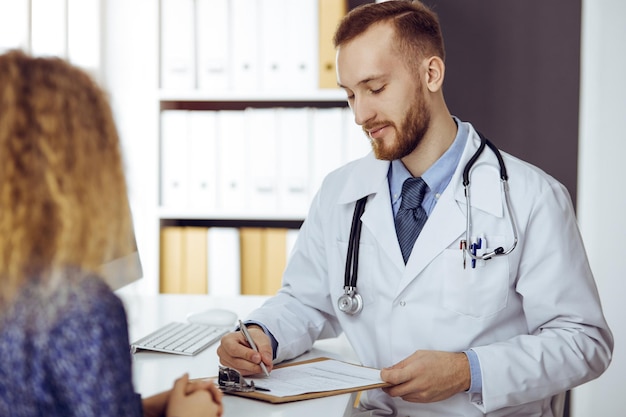 Freundlicher rotbärtiger Arzt und geduldige Frau diskutieren aktuelle Gesundheitsuntersuchung, während sie in sonniger Klinik sitzen. Medizin-Konzept.