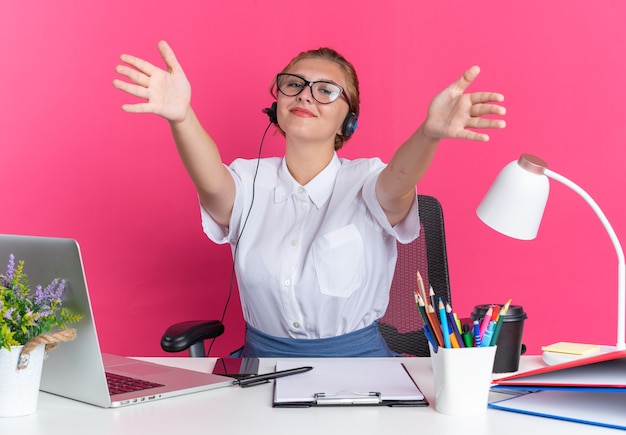 Freundliches junges blondes Callcenter-Mädchen mit Headset und Brille, das am Schreibtisch mit Arbeitswerkzeugen sitzt und eine Willkommensgeste macht