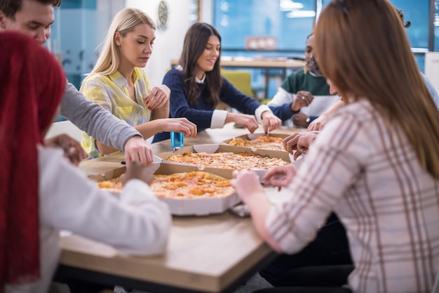 Freundliches, vielfältiges, multiethnisches Geschäftsteam, das gemeinsam Pizza im Startup-Büro isst, gemischtrassige Mitarbeitergruppe genießt die Pause, redet und lacht in der Mittagspause bei Meetings