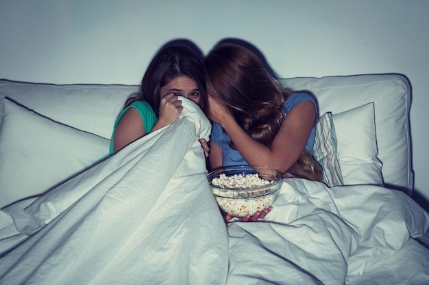 Freundschaft, Menschen, Pyjama-Party, Unterhaltung und Junk-Food-Konzept - verängstigte Freunde oder Mädchen im Teenageralter, die Popcorn essen und zu Hause Horrorfilme im Fernsehen ansehen