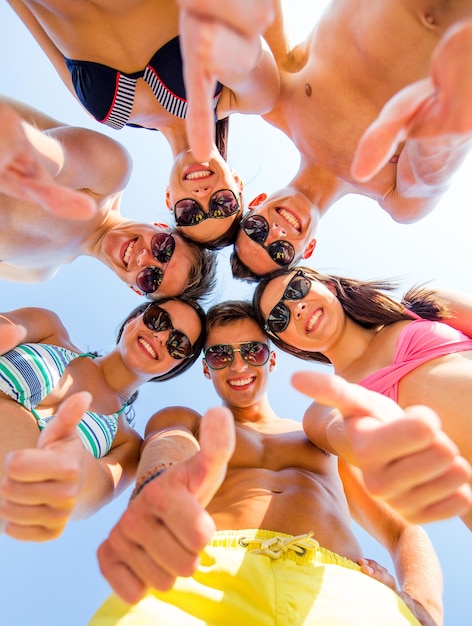 freundschaft, sommerferien, ferien, gesten und personenkonzept - gruppe lächelnder freunde, die badebekleidung tragen, die im kreis stehen und daumen hoch über blauem himmel zeigen