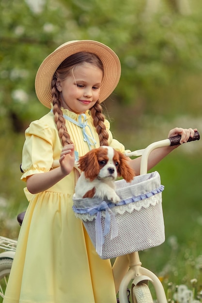 Freundschaft von Kindern und Haustieren Ein süßes Mädchen und ein kleiner Welpenkavalier fahren im Frühling Fahrrad in einem blühenden Garten