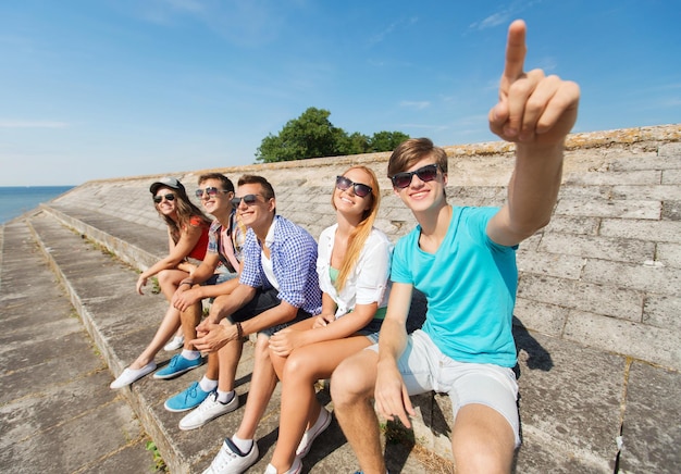 Freundschafts-, Freizeit-, Sommer-, Gesten- und Personenkonzept - eine Gruppe lächelnder Freunde, die auf der Straße der Stadt sitzen und mit dem Finger zeigen