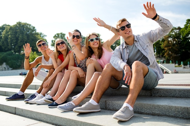 Freundschafts-, Freizeit-, Sommer-, Gesten- und Personenkonzept - eine Gruppe lächelnder Freunde, die auf der Straße der Stadt sitzen und mit den Händen winken