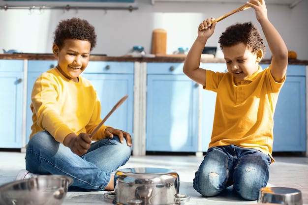 Freundschaftsziele. Freudige kleine Jungen sitzen mit gekreuzten Beinen auf dem Küchenboden und erzeugen Geräusche, indem sie auf Töpfen trommeln