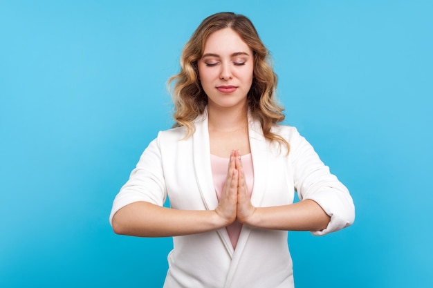 Frieden und Harmonie Stressbewältigung Porträt einer ruhigen Frau mit welligem Haar in weißer Jacke, die achtsames Yoga praktiziert, das Hände im Gebet hält und mit geschlossenen Augen meditiert Studioaufnahme blauem Hintergrund