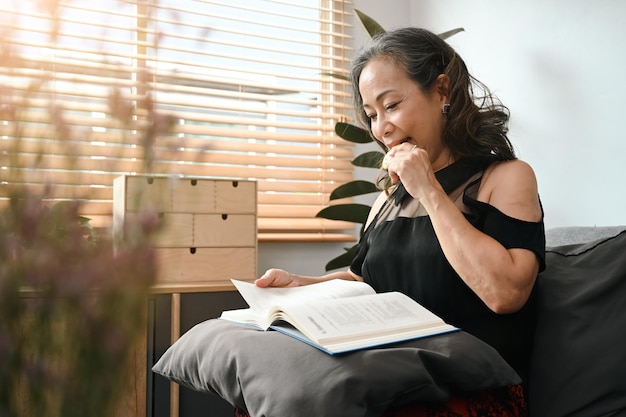 Friedliche Frau mittleren Alters, die Kekse isst und Buch auf der Couch zu Hause liest Freizeitaktivität positives Stimmungskonzept