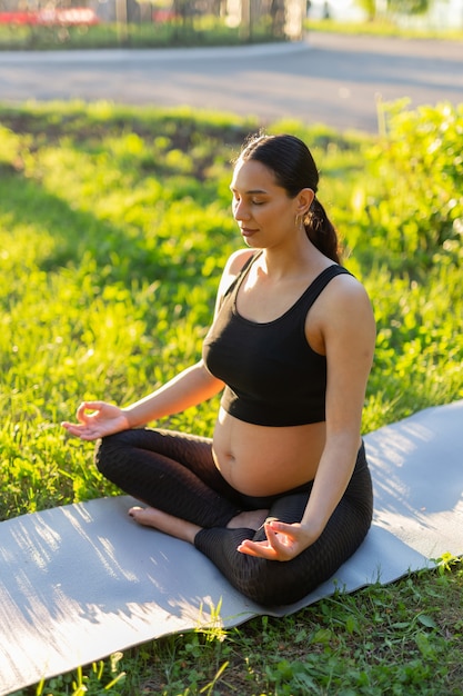 Friedliche junge positive schwangere Frau im Turnanzug macht Yoga und meditiert auf Matte auf grünem Gras am sonnigen warmen Sommertag sitzend. Konzept der Vorbereitung auf die Geburt und positive Einstellung