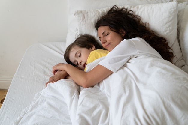 Friedliche liebevolle Mutter umarmt den Vorschulsohn, der zusammen mit dem Kind in einem gemütlichen Bett unter einer warmen Decke schläft