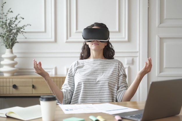 Friedliche Mitarbeiterin, die in einer Brille der virtuellen Realität meditiert und in Zen-Pose am Arbeitsplatz sitzt