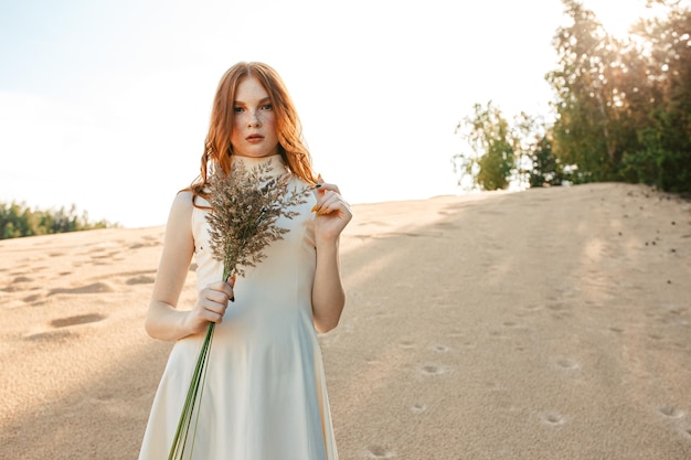 Friedliche rothaarige Frau mit Grasstrauß im Kleid, die im Sommer am Sandstrand steht