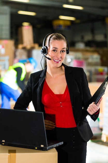 Friendly Woman, despachador o supervisor usando auriculares y computadora portátil en el almacén de la empresa de transporte,