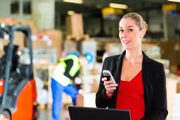 Foto friendly woman, despachador o supervisor usando un teléfono celular y una computadora portátil en el almacén de la empresa de transporte, una carretilla elevadora