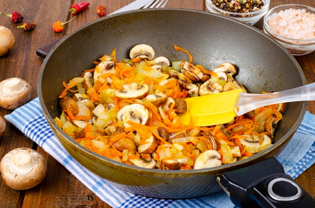 Frigideira com champignon de cogumelos fritos, cebola e cenoura na mesa de madeira.