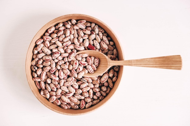 Frijoles secos crudos en un cuenco de madera con una cuchara sobre un fondo blanco.