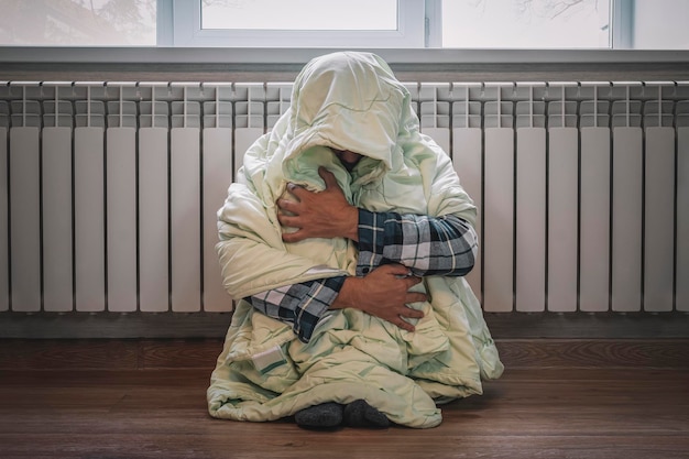 Frio em casa no inverno Homem congelando em sua casa no inverno por causa do termostato quebrado Cara jovem triste envolto em manta tremendo enquanto está sentado no chão no interior da sala de estar