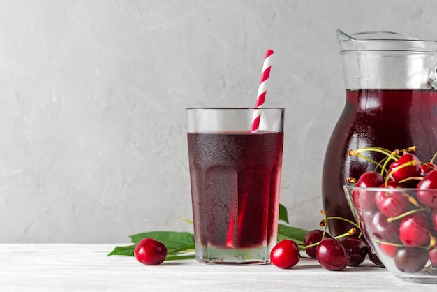 Frío jugo de cereza en un vaso con una pajita y jarra con bayas frescas en blanco. Refresco bebida de verano