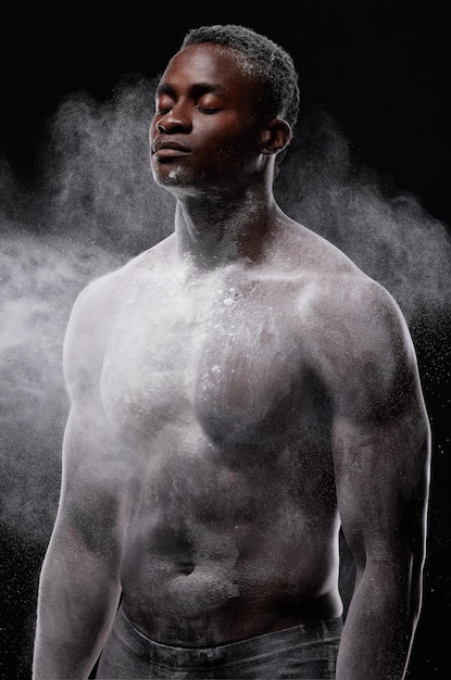 Frio. Tranquilo. Recogido. Confidente. Imagen artística de un joven musculoso posando con polvo blanco en el estudio sobre un fondo oscuro.