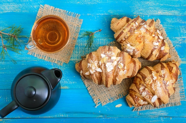 Frisch gebackene Croissants mit Mandelflocken, eine Teekanne und eine Tasse Tee auf blauem Holzhintergrund. Französisches Gebäck. Frühstück.