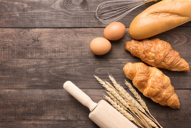 Frisch gebackene Hörnchen, Stangenbrot und Eier auf hölzernem Hintergrund