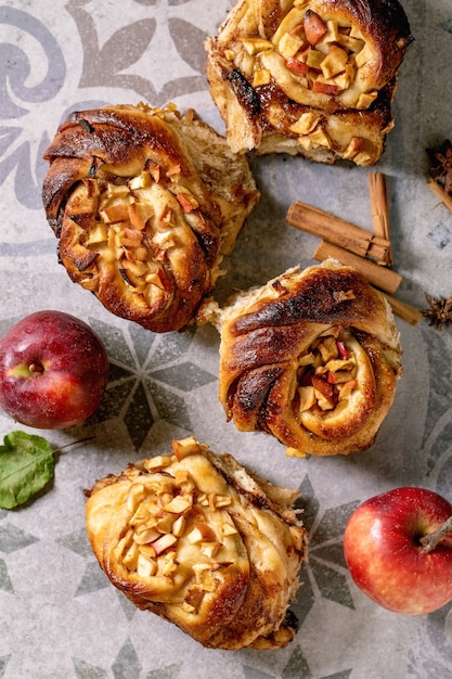 Frisch gebackener süßer Apfel klebrige Zimtbrötchen mit Gartenäpfeln und Zimtstangen auf verziertem Keramikfliesenhintergrund. Traditionelle Hausbäckerei