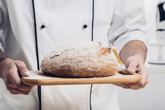 Frisch gebackenes Brot auf einem Holzbrett in den Händen eines Bäckers. Sanftes Licht