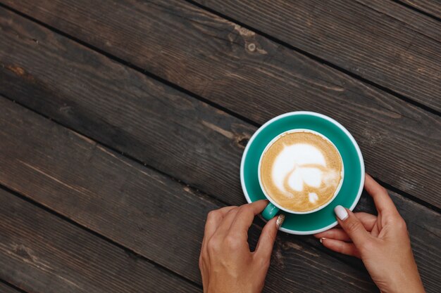 Frisch gebrühter heißer Kaffee mit schöner Latte Art in Herzform in türkisfarbenem Tassenhalter von einer Frau in Händen