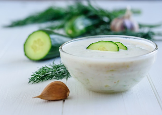 Frisch gemachtes Tzatziki in einer Glasschüssel mit Gurke auf einem weißen Holztisch.