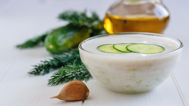 Frisch gemachtes Tzatziki in einer Glasschüssel mit Knoblauch und Petersilie auf einem weißen Holztisch.