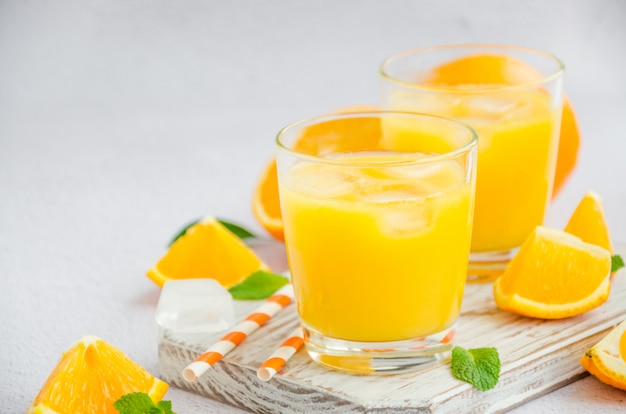 Frisch gepresster Orangensaft mit Eis in einem Glas mit einem Strohhalm auf einem Holzbrett auf einer hellen Oberfläche mit frischen Orangen. Horizontale Ausrichtung, Kopierraum.