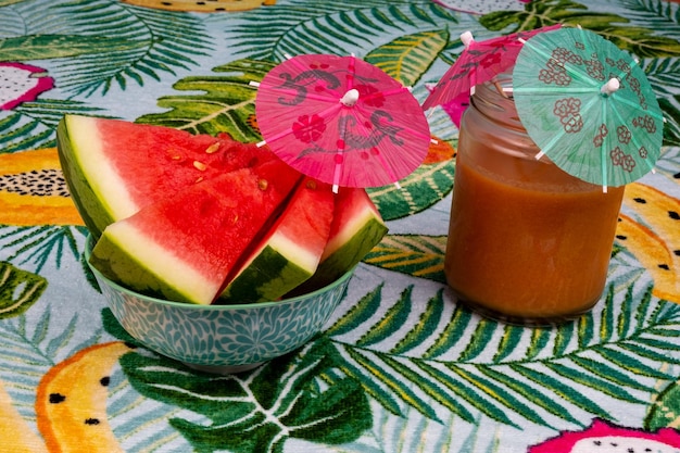 Frisch geschnittene Wassermelone mit kleinem Regenschirm, ein Glas Gazpacho auf einem Tisch mit Palmen.