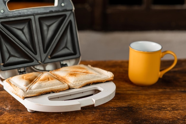 Frisch zubereitete geröstete Sandwiches in einem Sandwichmaker auf Holzhintergrund. Daneben befindet sich eine Tasse Morgenfrühstückskonzept