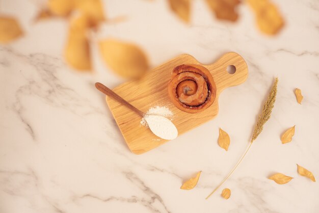 Frische Bäckerei, frisches Zimtbrötchen mit dem Löffel voll vom Mehl auf hölzernem Brett auf weißer Marmoroberfläche. Leckeres leckeres Dessert, französisches Frühstück. Ansicht von oben