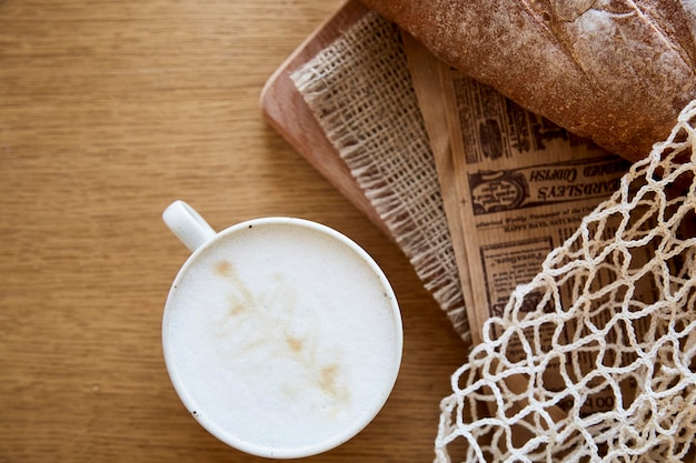 Frische Baguette-Zeitung und Cappuccino auf Holztisch im französischen Eco-Mesh-Frühstück